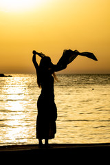 Beautiful female silhouette of unrecognizable slim tall woman with silk scarf at sunset in front of golden Ionian Sea water. Colorful dusk as seen from Ksamil, Albania, late spring