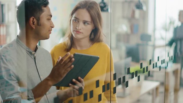 Portrait of Handsome Indian Man Showing Touch Screen Digital Tablet Computer To His Beautiful Female Team Member, They Discuss Project Details. Successful Young People Working in Diverse Office