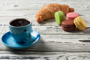 One full blue ceramic cup of fresh black coffee near whole baked croissant and various macaroons on old white rustic wooden table