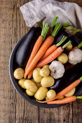 Raw  Potatoes Carrot Garlic and Eggplant on Black Plate Variety of Raw Vegetables ready to Cook Fresh Natural Vegetables Organic Healthy Food Olive Oil Pepper Pot Wooden Background Vertical Top View
