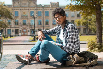 Obraz na płótnie Canvas A black African American student in glasses with books sits near a college.