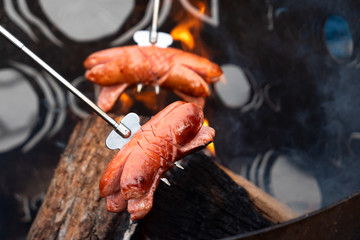 Two sausages on metal forks over outdoor camp fire.