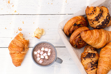 Continental breakfast with hot chocolate with marshmallows and basket of pastries. Half eaten on white wood from above.