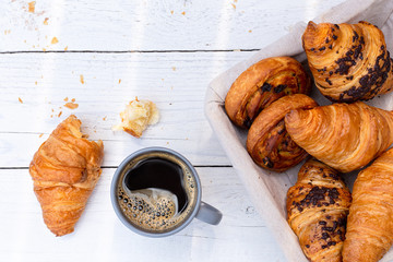 Continental breakfast with black coffee and basket of pastries. Half eaten on white wood from above.