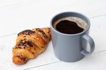 Black coffee in a blue-grey ceramic mug next to chocolate croissant isolated on white painted wood.