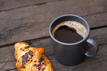 Black coffee in a blue-grey ceramic mug next to chocolate croissant isolated on rustic dark brown wood table.