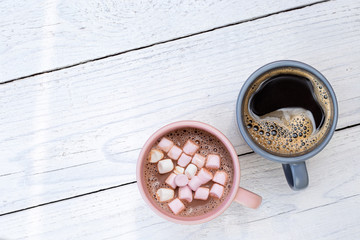 Mug of hot chocolate with small marshmallows next to a mug of black coffee isolated on white painted wood from above. Space for text.