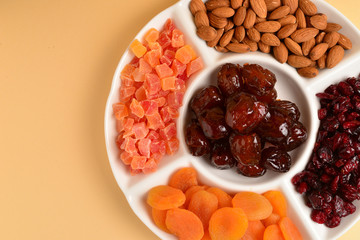Mix of dried fruits and nuts on a white plate. Apricot, almond, raisin, dates fruit. On a brown background. Space for text or design.