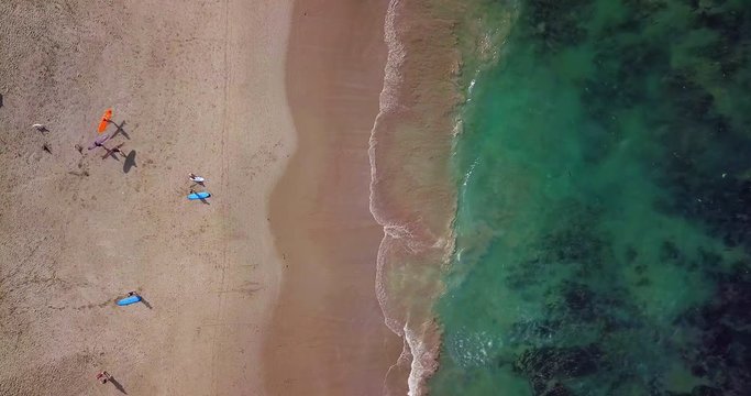 Timelapse. High aerial top view of tiny surfers on sand beach. Small toy figures go into water. Sea waves on beautiful beach aerial view drone shot. Bird's eye view of ocean waves crashing at beach.