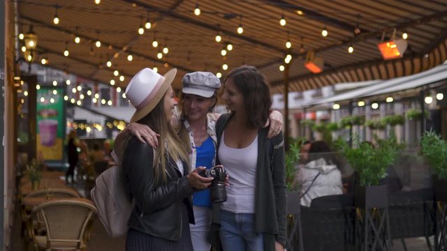 Two traveling girl friends checking photos on dslr camera and their friend hugs them in Riga city - Travel tourism concept after transfer from airport - Wearing hats and jeans