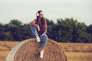 Naklejka na ściany i meble Beautiful girl sitting on a haystack, listening to music , enjoying sunset.