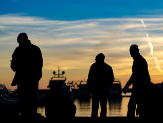 Fototapeta na wymiar Dark silhouettes of a group of people on the sea pier of the yacht marina against the sunset, silhouettes of yachts and blue sky