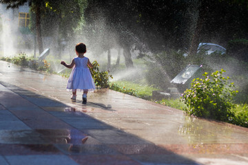 Cute baby girl in a beautiful white gown walking across a sidewalk at a lawn with running water sprinklers in a sunny day. Childhood and Happiness concept.