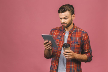 Portrait of confident young handsome man in casual with cup of coffee working on digital tablet isolated on pink background in studio.