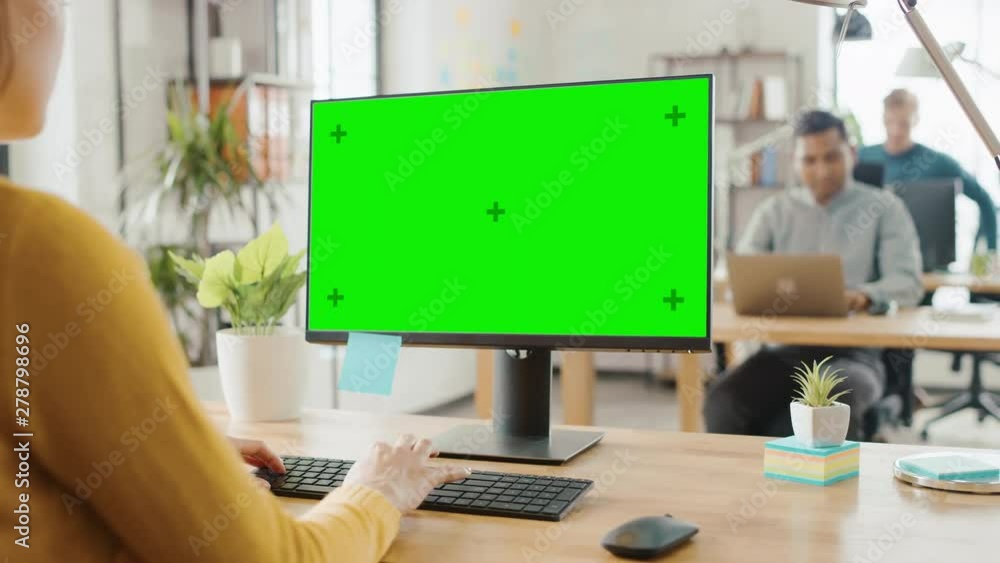 Poster over the shoulder: creative young woman sitting at her desk using desktop computer with mock-up gree