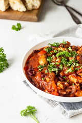 Meat stew with red beans, bell pepper and onion in tomato sauce in a white plate over light grey slate or stone background. 