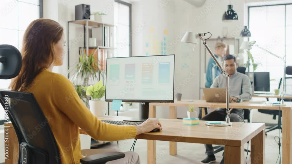 Poster Over the Shoulder: Female Mobile Software Developer Sitting at Her Desk Using Desktop Computer with Screen Showing Smartphone Application UI / UX. Office With Diverse Team of Professionals Working