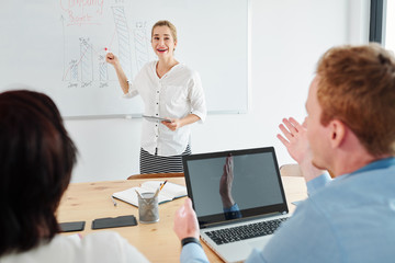 Happy young businesswoman standing and pointing at whiteboard with graphics and explaining it to her colleagues during presentation at office