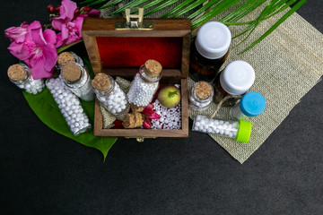 Top view of homeopathic medicine bottles, scattered pills with pink flower and wild fruit in wooden old box, burlap and green leaf on dark background. Natural Homeopathy