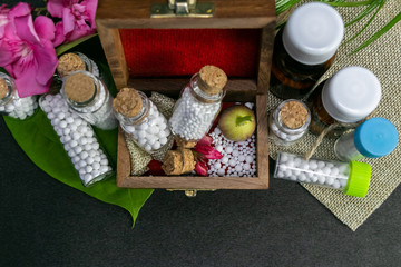 Top view of homeopathy pills in vintage bottles, scattered pills with pink flower, cork and wild fruit in wooden old box, burlap and green leaf on dark background. Natural Homeopathic concept