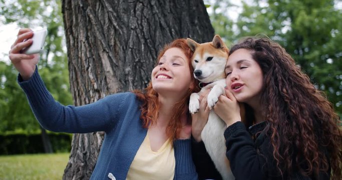 Loving dog owners attractive curly-haired girls are taking selfie with pet in park using smartphone camera hugging and strokign animal. People and emotions concept.