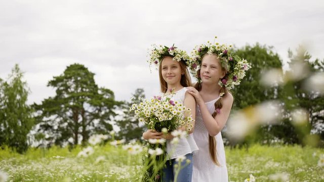Two girl friends in wreath with flowers bouquet standing on blooming field. Teenager girls posing on flowering field at sunny summer day