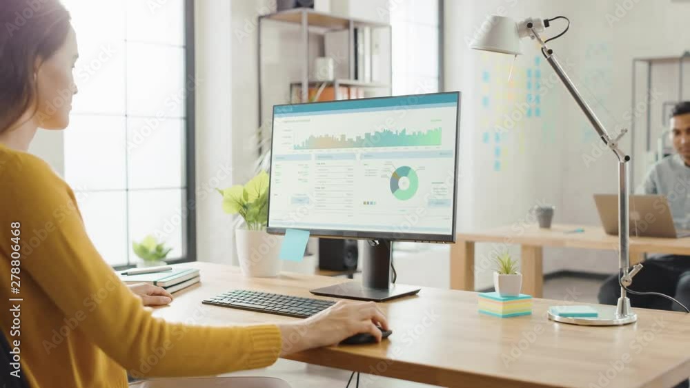 Poster Over the Shoulder: Creative Young Woman Sitting at Her Desk Using Desktop Computer with Screen Showing Project Management, Statistics and Graphs. Office with Diverse Team of Professionals Working