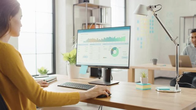 Over the Shoulder: Creative Young Woman Sitting at Her Desk Using Desktop Computer with Screen Showing Project Management, Statistics and Graphs. Office with Diverse Team of Professionals Working