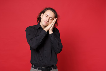 Young curly long haired man in black shirt posing isolated on red wall background studio portrait. People sincere emotions lifestyle concept. Mock up copy space. sleep with folded hands under cheek.