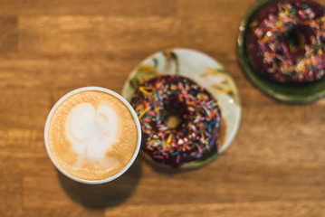 Coffee with a drawn heart and milk on a wooden table in a coffee shop. two chocolate donuts with scattering on the table next to the coffee