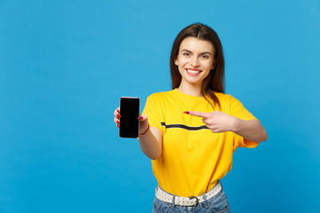 Portrait of cheerful young woman in vivid casual clothes pointing index finger on mobile phone with blank empty screen isolated on bright blue background. People lifestyle concept. Mock up copy space.
