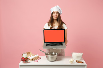 Chef cook confectioner or baker in white t-shirt, toque chefs hat cooking Christmas cake or cupcake at table hold pc laptop with blank empty screen isolated on pink pastel background. Mock up concept.
