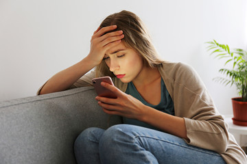 Portrait of beautiful young woman with depressed facial expression sitting on grey textile couch...
