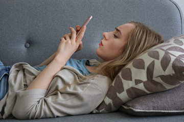 Fototapeta na wymiar Portrait of beautiful young woman with depressed facial expression sitting on grey textile couch holding her phone. Cyber bullying victim concept. Sad female in her room. Background, copy space.