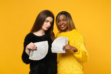 Two fun young women friends european and african in black yellow clothes holding fan of cash money in dollar banknotes isolated on orange wall background. People lifestyle concept. Mock up copy space.