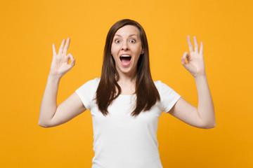 Portrait of amazed young woman in white casual clothes keeping mouth open, showing OK gesture isolated on bright yellow orange wall background in studio. People lifestyle concept. Mock up copy space.