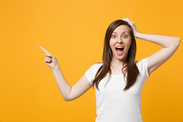 Amazed young woman in white casual clothes keeping mouth open, pointing index finger aside, putting hnd on head isolated on yellow orange wall background. People lifestyle concept. Mock up copy space.