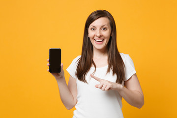 Naklejka premium Smiling young girl in white casual clothes pointing index finger on mobile phone with blank empty screen isolated on bright yellow orange wall background. People lifestyle concept. Mock up copy space.