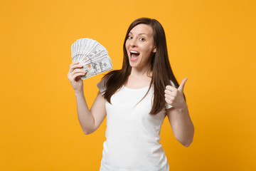Surprised young girl in white clothes keeping mouth open showing thumb up hold fan of cash money in...