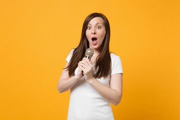 Portrait of shocked amazed young woman in white casual clothes looking camera sing song in microphone isolated on yellow orange wall background in studio. People lifestyle concept. Mock up copy space.