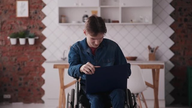 Young and disabled man wearing in denim clothes sitting in special comfort wheelchair and spending weekend using laptop. Male relaxing or working at home with bright light room in modern interior flat