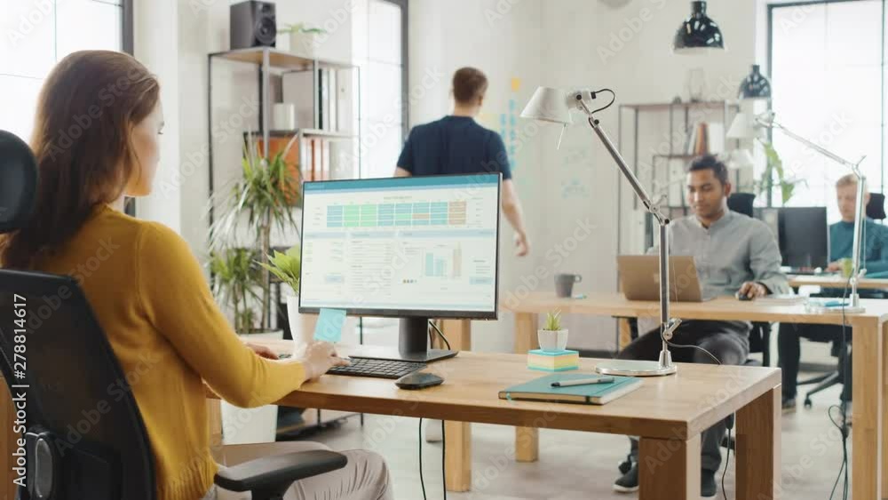 Poster Over the Shoulder: Creative Young Woman Sitting at Her Desk Using Desktop Computer with Screen Showing Project Management, Statistics and Graphs. Office with Diverse Team of Professionals Working