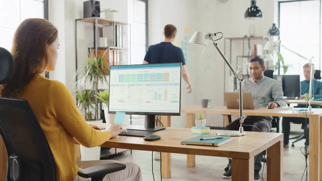 Over the Shoulder: Creative Young Woman Sitting at Her Desk Using Desktop Computer with Screen Showing Project Management, Statistics and Graphs. Office with Diverse Team of Professionals Working