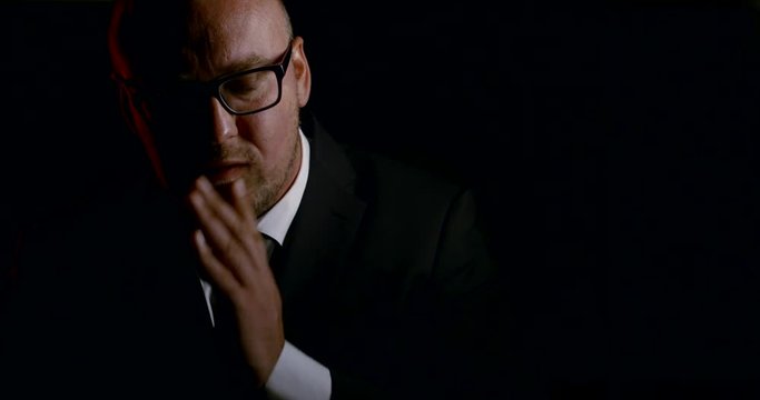 Portrait of a bald sad man in glasses and a business suit with a tie, it is on a black background. He is holding his head and face, holding and thinking of suicide.