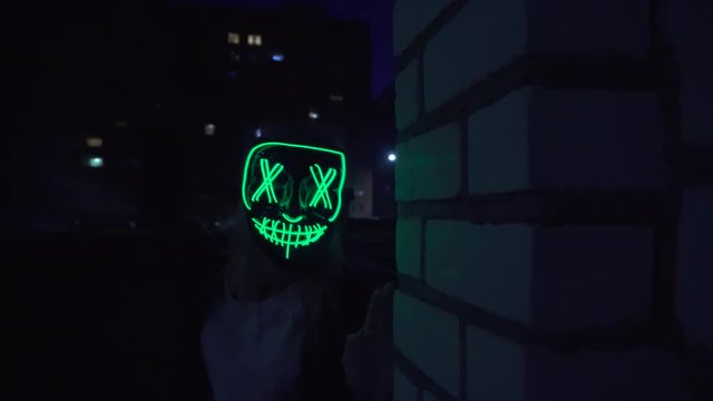 I pop up when the rave starts. Brick wall. A man wears an exotic LED mask. The mask is lit, glowing, street, night, city, light.