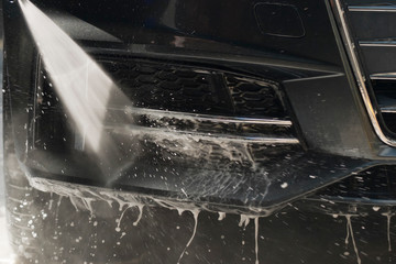 Car washing with high pressure water jet. Water and foam under pressure flies toward to the front bumper of the car body. Close-up view