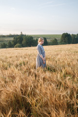 Naklejka na ściany i meble young girl in blue dress with collected hair looks at camera in field of ears at sunrise. Green boscage in background