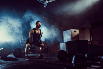 Muscular attractive caucasian bearded tasttoed man lifting kettlebell in a gym. Weight plates, dumbbell and tires in smoke background.