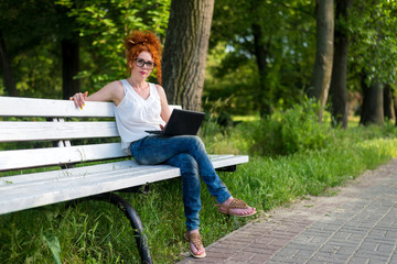 Fototapeta premium Red-haired female freelancer working on a laptop while sitting on a park bench.