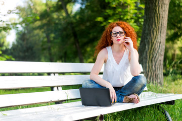 Red-haired female freelancer working on a laptop while sitting on a park bench.